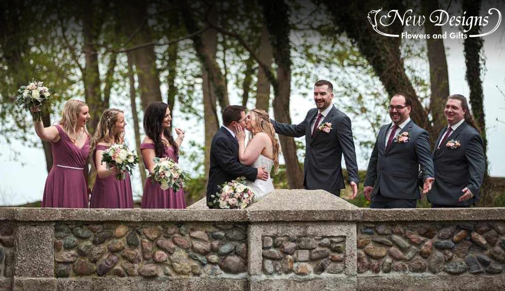 A wedding party cheers on the bride and groom on a cobblestone bridge as they kiss for the camera, the bouquets and pocket square flowers in gorgeous shades of white and pink